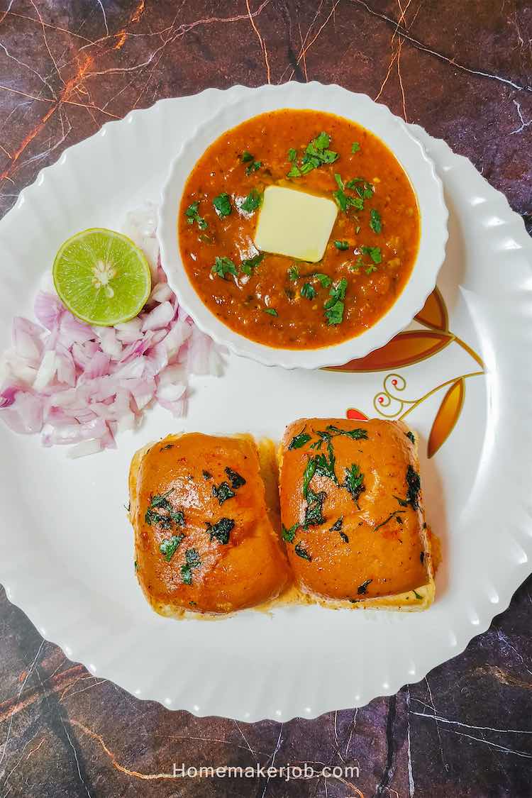 Pav bhaji served in a white plate with two pav buns, bhaji in a white bowl with butter cube on a top, and chopped onion along with lemon besides the bowl
