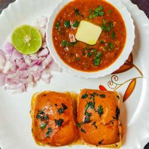 Pav bhaji served in a white plate with two pav buns, bhaji in a white bowl with butter cube on a top, and chopped onion along with lemon besides the bowl