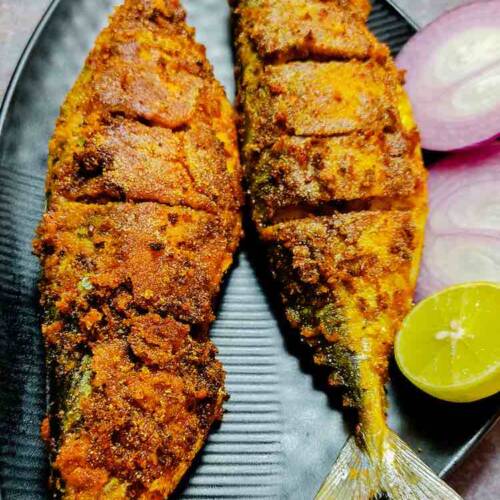 Two full pieces of ready spicy fried bangada (indian mackerel) fish garnished with onions and lemon served in a black dish