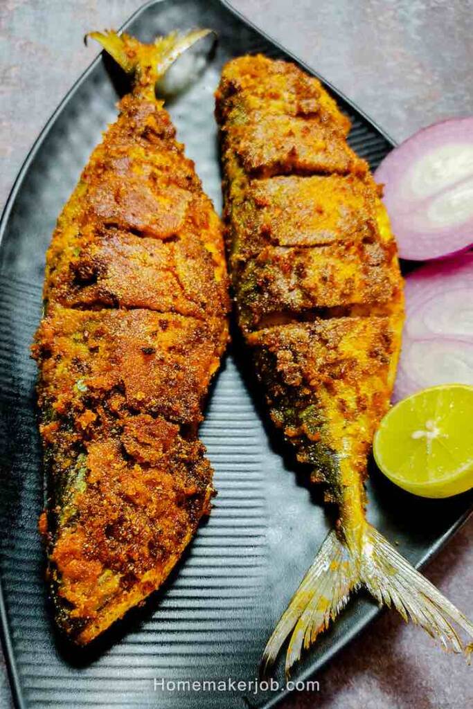 Two full pieces of ready spicy fried bangada (indian mackerel) fish garnished with onions and lemon served in a black dish