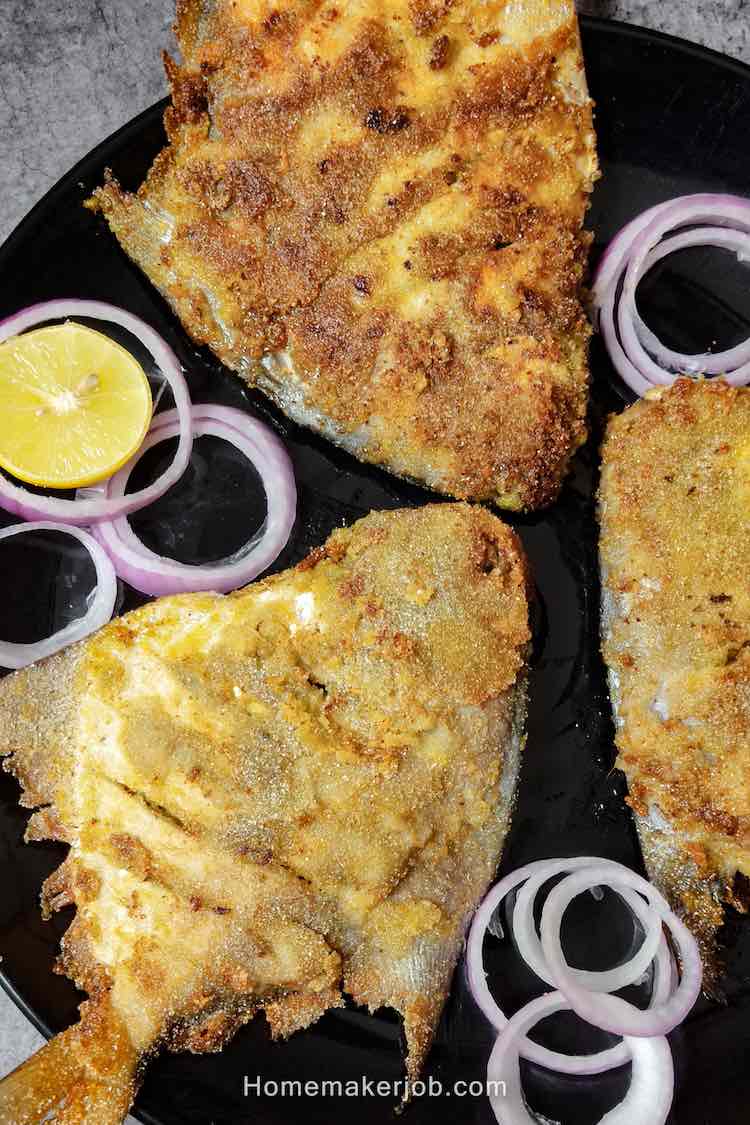 Close up photo of ready pieces of stuffed pomfret green masala fry served in a bliack dish garnished with onions and lemon wedges by homemakerjob