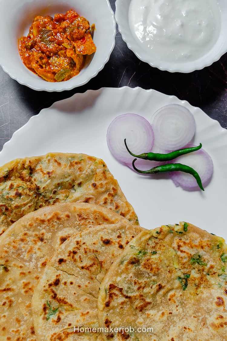 close up photo of four punjabi aloo paratha breads served in a white dish garnished with sliced onions and two green chilies accompanied by mango pickle and curd in two separate bowls