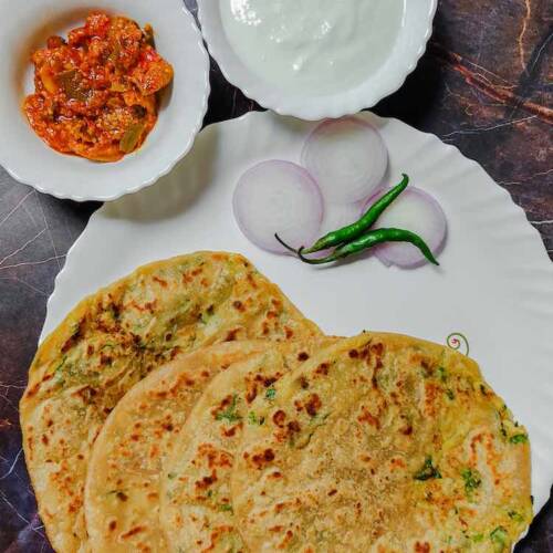 4 punjabi aloo paratha breads served in a white dish garnished with sliced onions and two green chilies accompanied by mango pickle and curd in two separate bowls