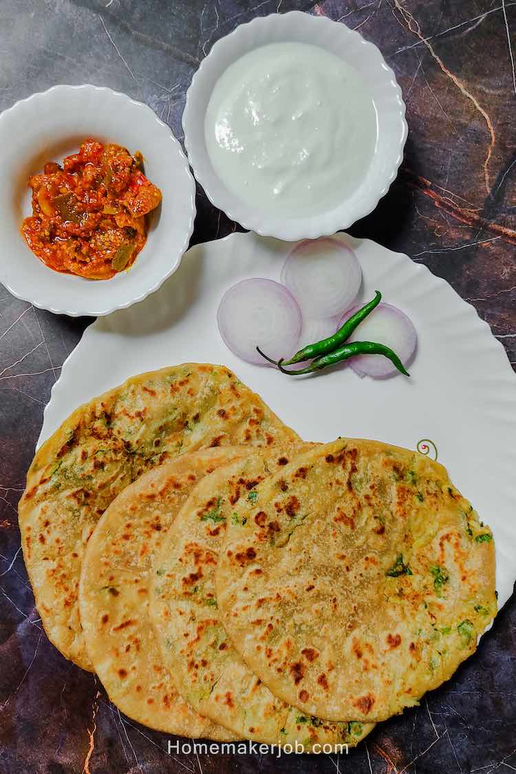 4 punjabi aloo paratha breads served in a white dish garnished with sliced onions and two green chilies accompanied by mango pickle and curd in two separate bowls