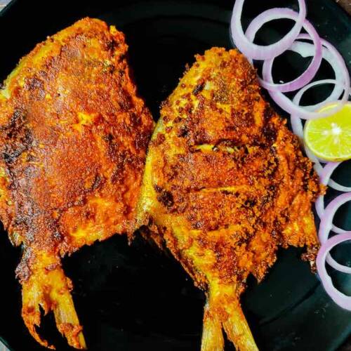 Photo of two full pomfret fish fried in roasted spices, served on a black plate and garnished with onions and lemon wedges by homemakerjob.com