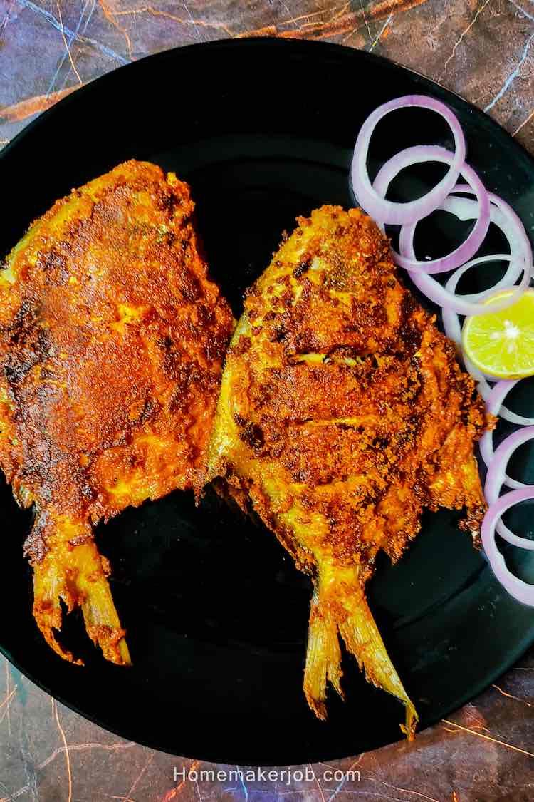 Photo of two full pomfret fish fried in roasted spices, served on a black plate and garnished with onions and lemon wedges by homemakerjob.com
