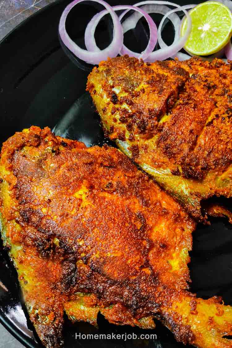 Close-up Photo of two full pomfret fish fried in roasted spices, served on a black plate and garnished with onions and lemon wedges by homemakerjob.com