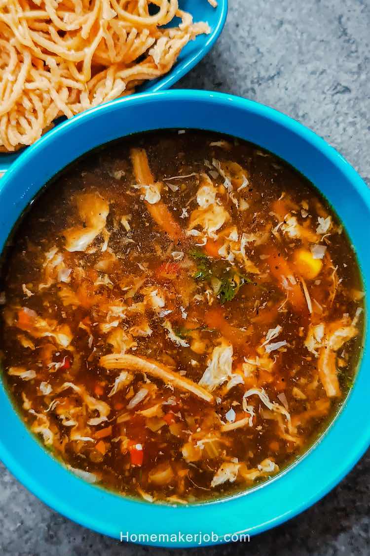 Close up photo of hot chicken manchow soup served in a blue bowl accompanied with dry fried noodles in a blue bowl on a table top, by homemakerjob.com