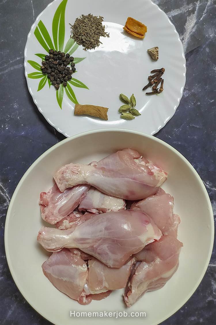 Two white dishes arranged one below another with spices arranged in round manner in top dish and raw chicken in bottom dish