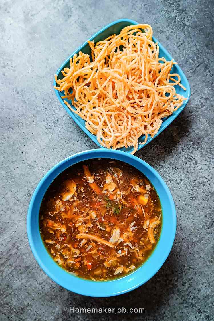 Photo of hot chicken manchow soup served in a blue bowl accompanied with dry fried noodles in a blue bowl on a table top, by homemakerjob.com