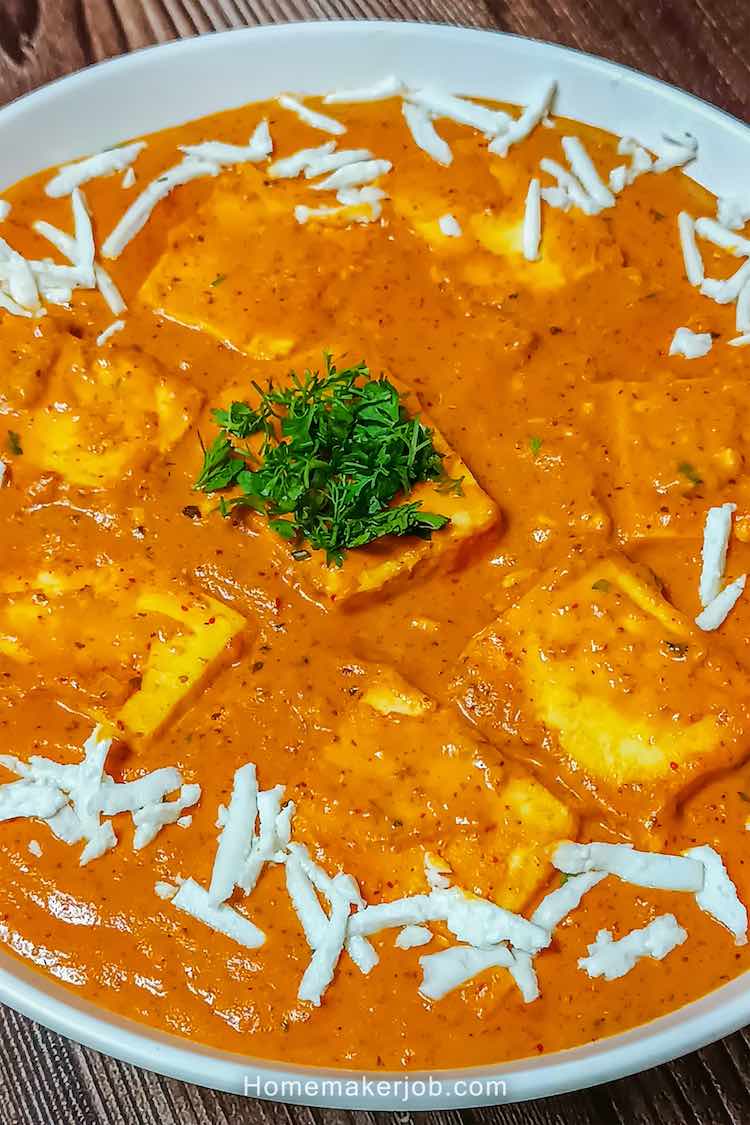 Close up photo of paneer lababdar served hot in a white dish on a table, garnished with chopped coriander in the middle and grated cottage cheese around the edges, a recipe by homemakerjob