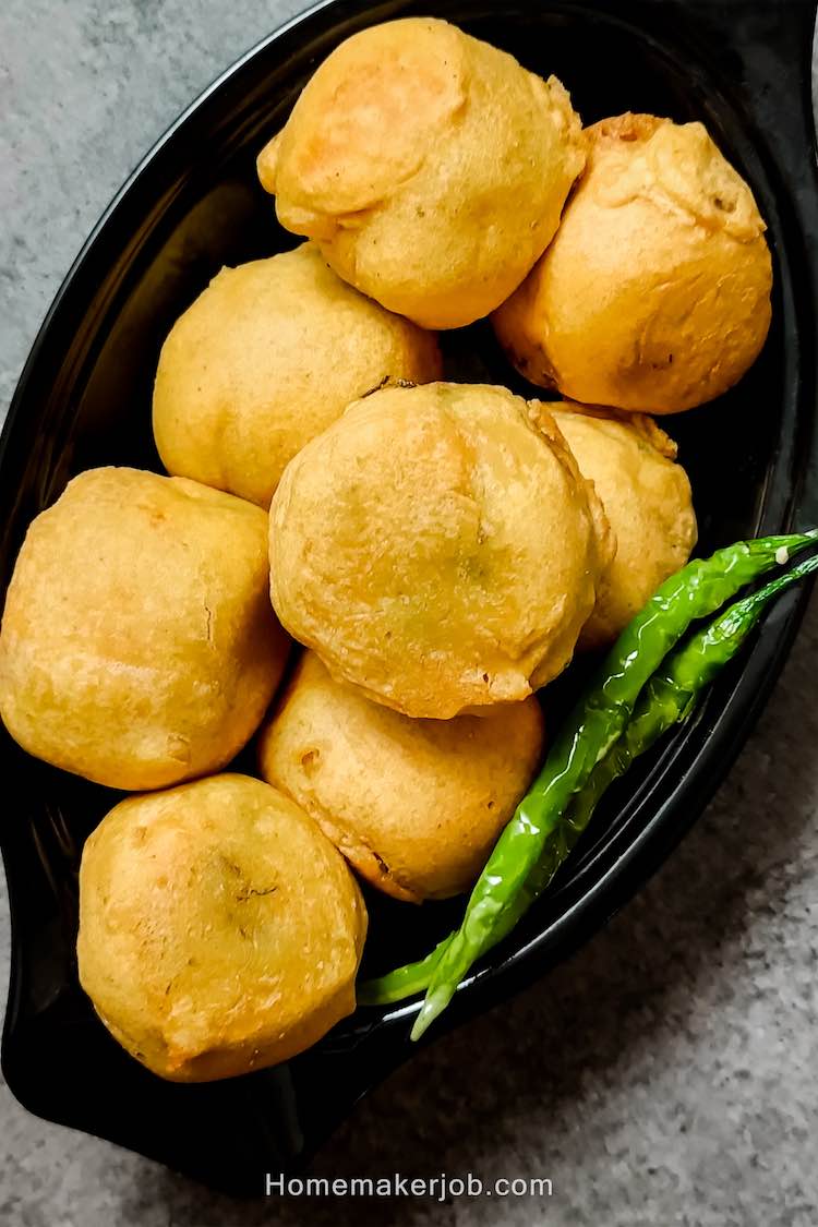 Close up photo of ready batata vada garnished with green chilies and served hot in a black dish on a table, by homemakerjob