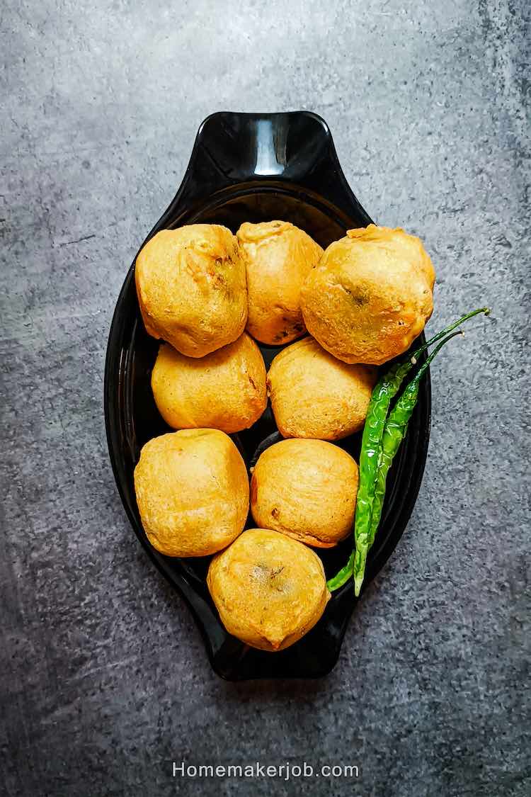 Photo of ready batata vada garnished with green chilies and served hot in a black dish on a table, by homemakerjob