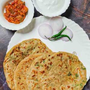 4 punjabi aloo paratha breads served in a white dish garnished with sliced onions and two green chilies accompanied by mango pickle and curd in two separate bowls