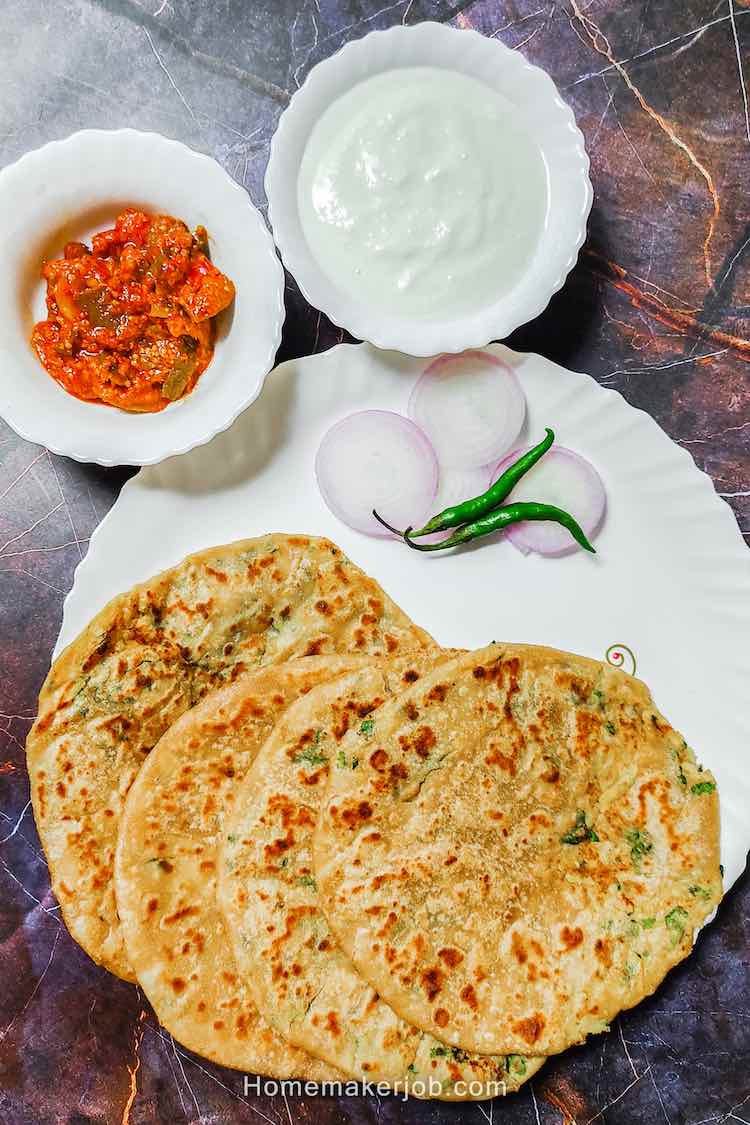 4 punjabi aloo paratha breads served in a white dish garnished with sliced onions and two green chilies accompanied by mango pickle and curd in two separate bowls