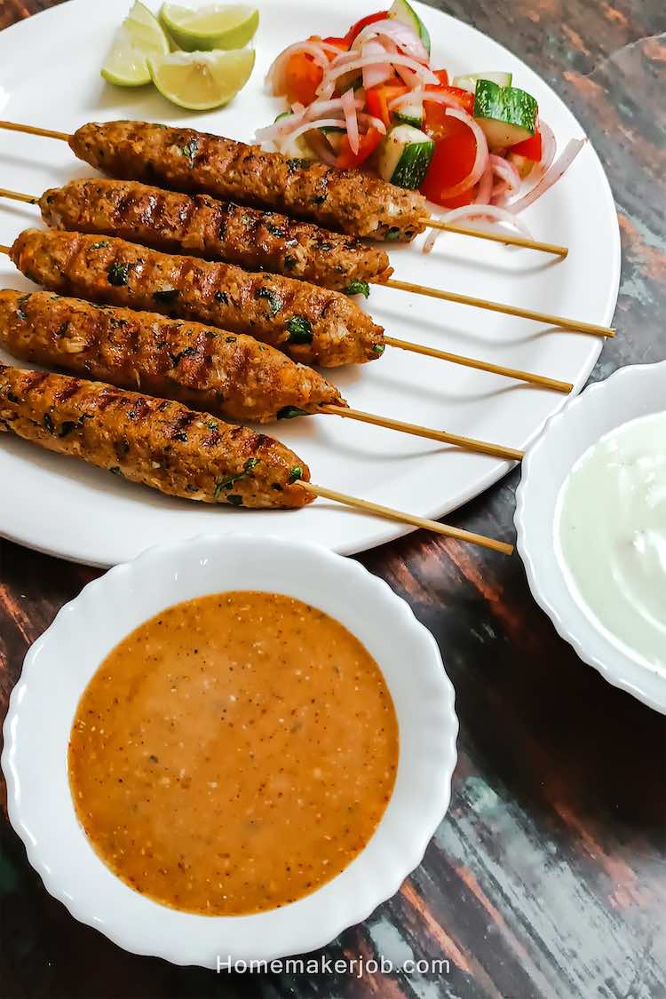 Top view close-up photo of chicken kofta kebabs (a.k.a. kabobs or kababs) on skewers with accompaniments as sauces, in a white dish on a table, a recipe by homemakerjob.com