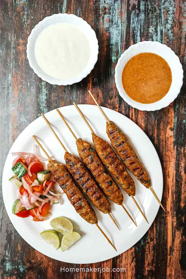 Top view photo of chicken kofta kebabs (a.k.a. kabobs or kababs) on skewers with accompaniments as cut lemons and salad, in a white dish on a table, a recipe by homemakerjob.com