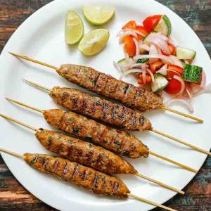 Top view photo of chicken kofta kebabs (a.k.a. kabobs or kababs) on skewers with accompaniments as cut lemons and salad, in a white dish on a table, a recipe by homemakerjob.com